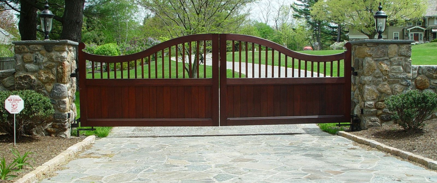 A custom mahogany wood driveway gate with a rich finish, featuring a modern arched design in a residential drivewa
